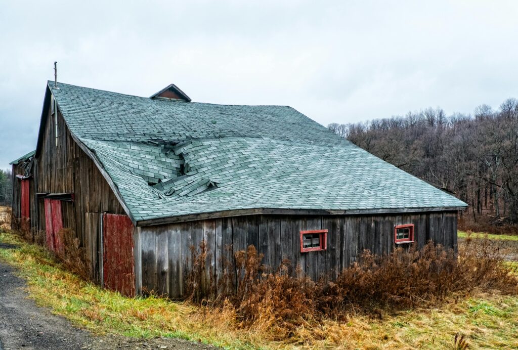 Post-Winter Home Inspection Checklist: Where to Start A weathered wooden barn with a damaged roof in a rural New York landscape.